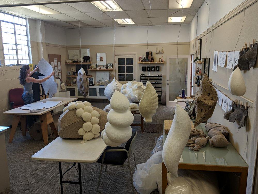 Artist Stephanie Metz stands in her studio examining a felt shape while the room is filled with felt and wool creations.