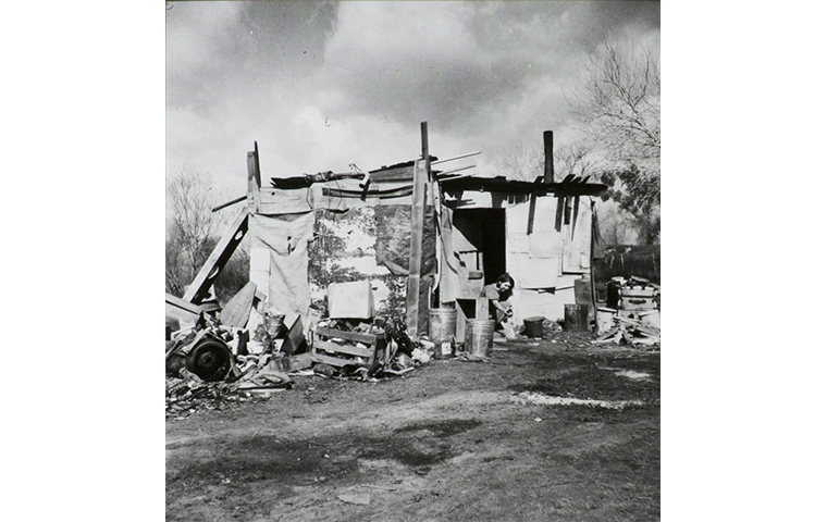 Black and white photograph of shack made of boards, tar paper and other salvaged materials. Debris and trash around building, a figure sits in the doorway. Trees and fields in the background.