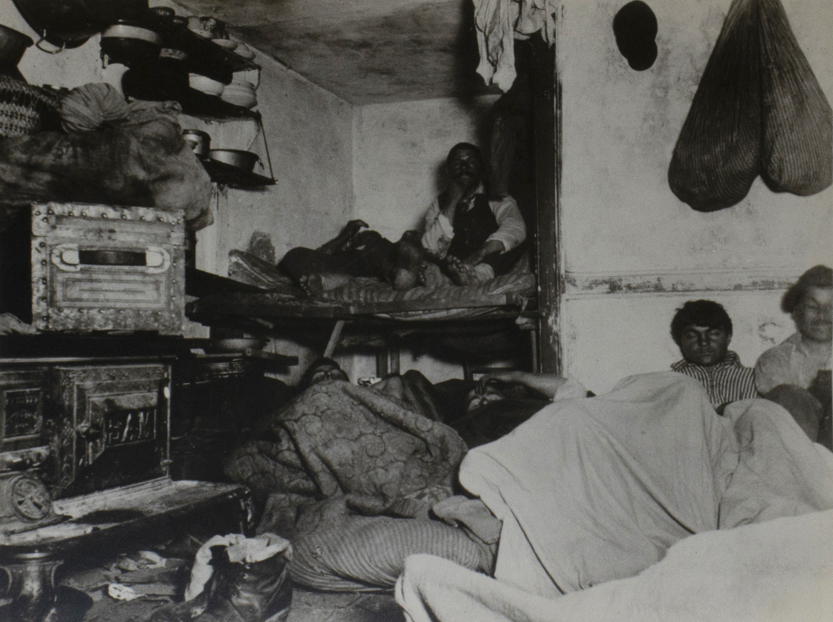 Black and white photograph of a cramped room in a tenement house. Two people to a bed. Two sacks hanging on the wall, on the right, over the heads of two men beneath a blanket. Stove and pots and a trunk on the left side.