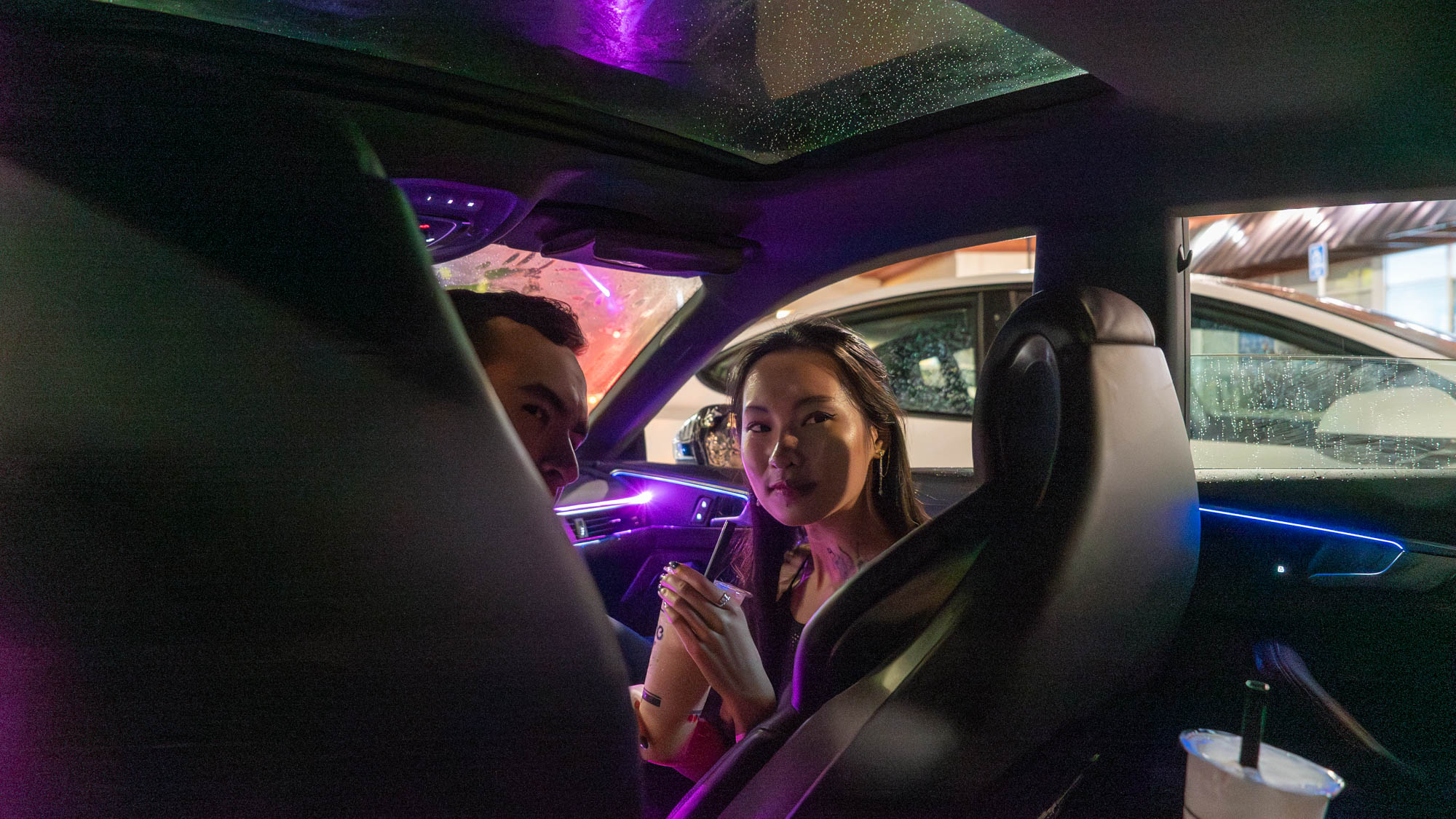 Photo of two Asian American young adults inside their car near a boba shop.