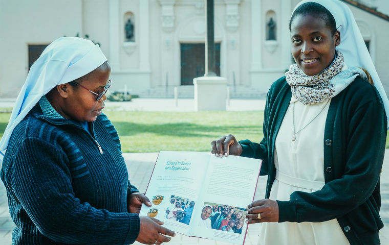 Two ACWECA Sisters holding a booklet in front of Mission Church image link to story