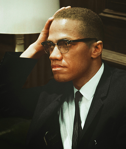 Malcolm X seated with hand resting on head, lamp in the background