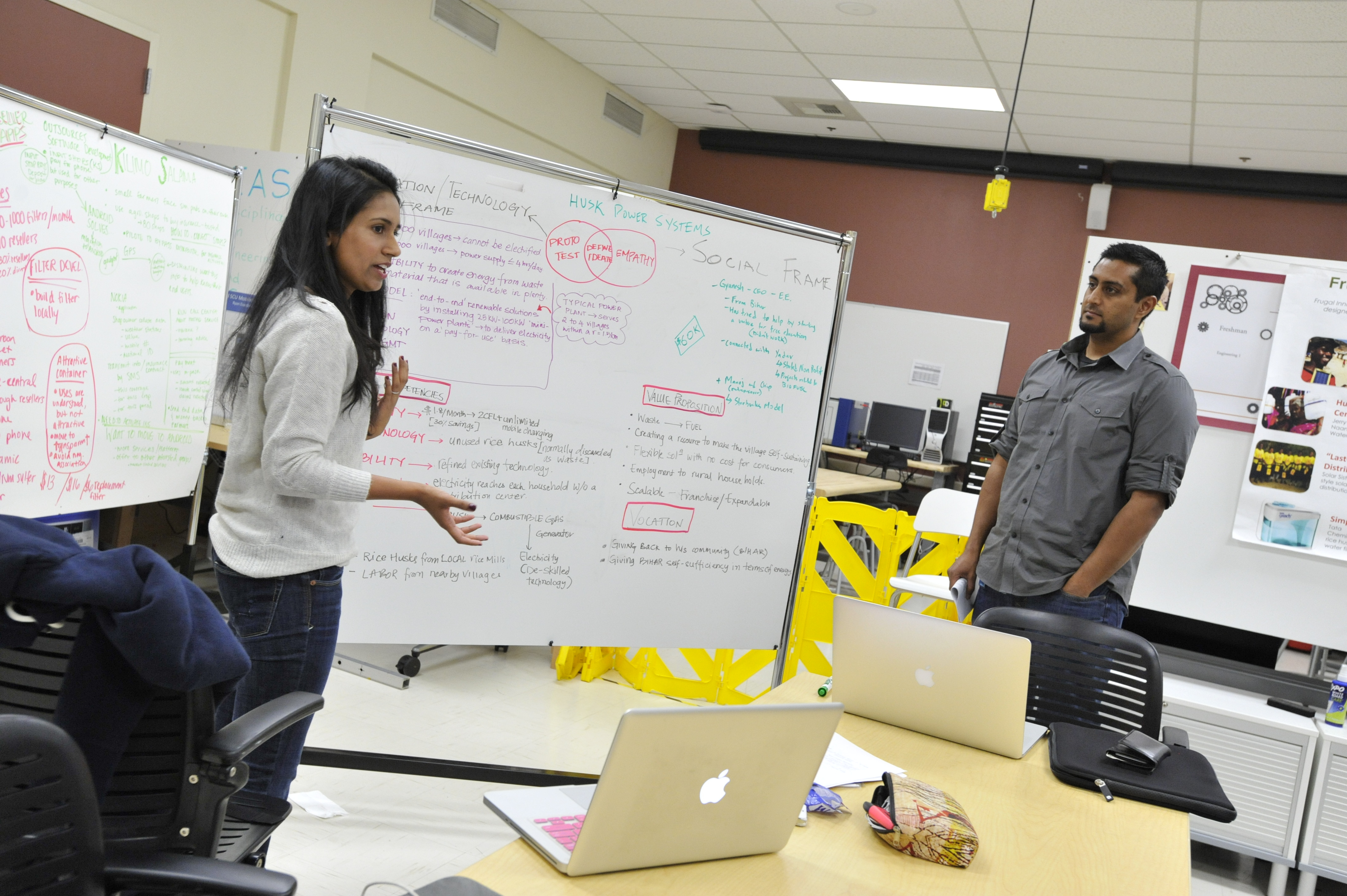 Two students stand in front of a white board with brainstorming work on it, presenting to colleagues.