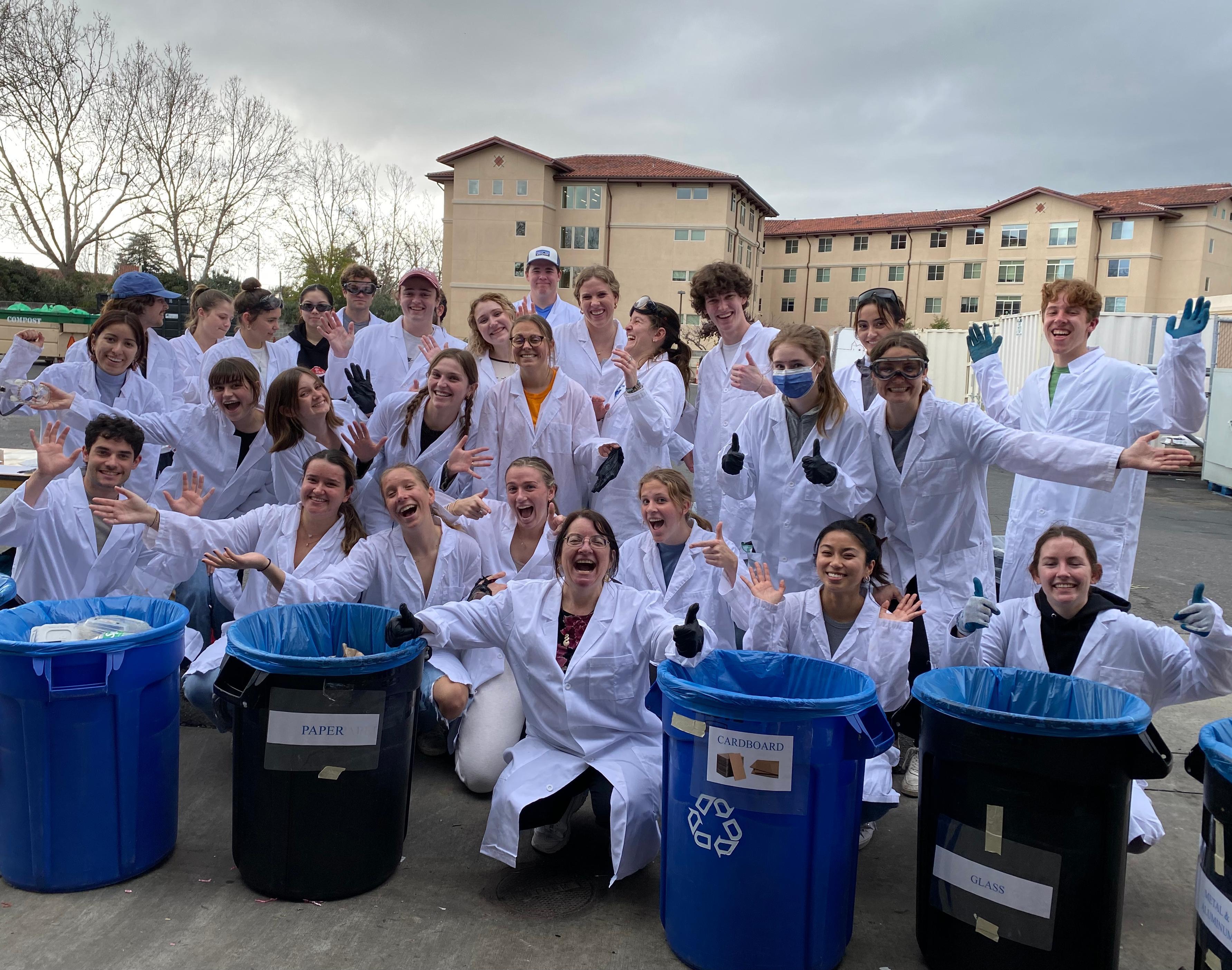 A group of students wearing lab coats by trash bins during a waste characterization event 