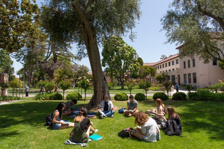 A faculty member and students sit outside in a circle discussing and taking notes