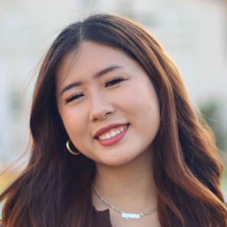 A person smiling outdoors with buildings in the background.