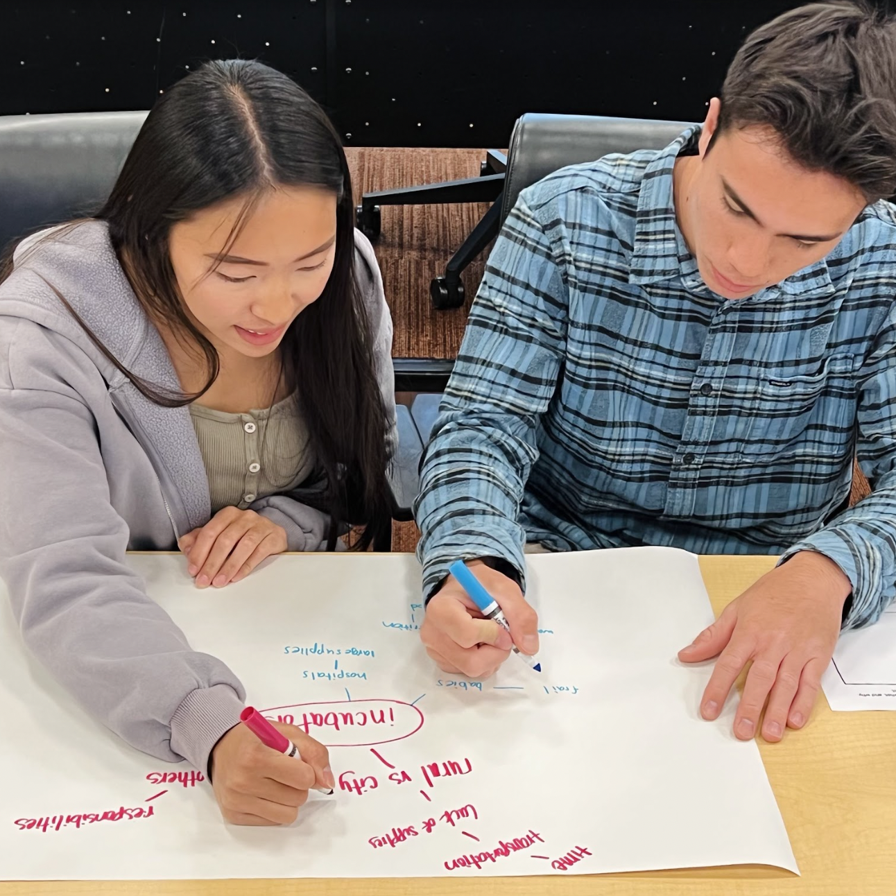 Two people writing on a large white sheet at a table.