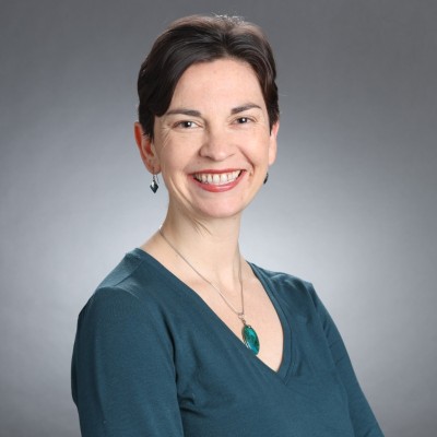 A smiling person wearing a teal top against a gray background.