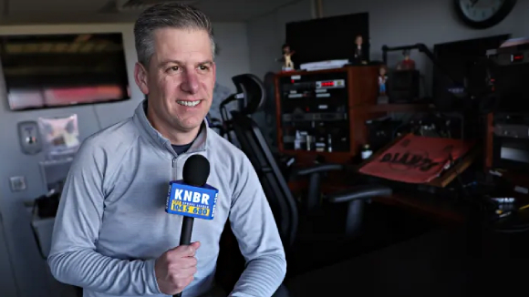 A man in athleticwear holds a microphone labeled KNBR.