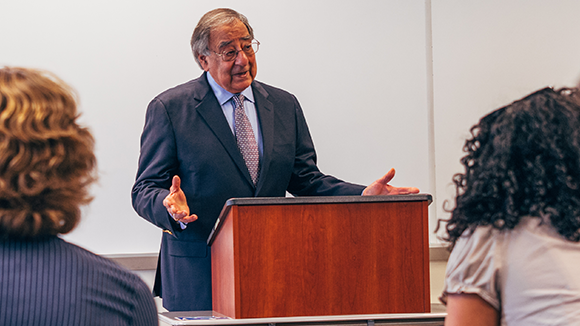 A man in a suit and tie speaks from a podium to a classroom full of students.