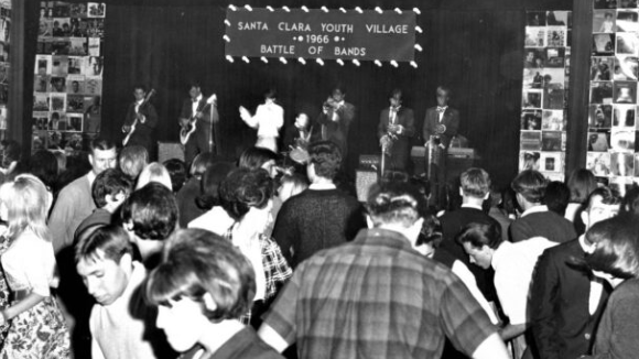 Young men and women in 1960s clothing dancing in front of a stage with a band. A sign over the band reads Santa Clara Youth Village. 1966. Battle of Bands.
