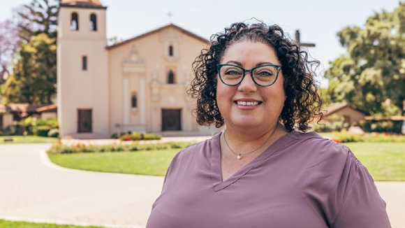 A smiling woman in glasses stands in front of Santa Clara Universitys Mission Church.