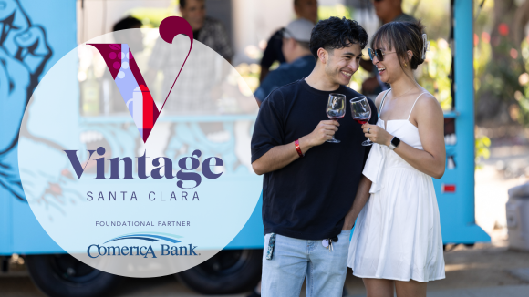 A young couple in front of a food truck stand close together, holding wine glasses in a cheers motion. Beside them is the Vintage Santa Clara logo and text underneath that reads Foundational Partner just over the Comerica Bank logo.
