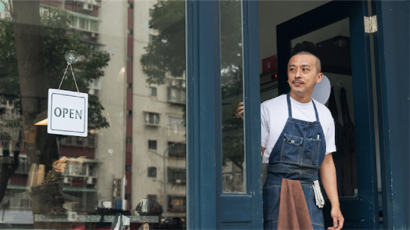 A man in a denim apron with a towel in the pocket looks out of a storefront. A sign in the window reads Open.