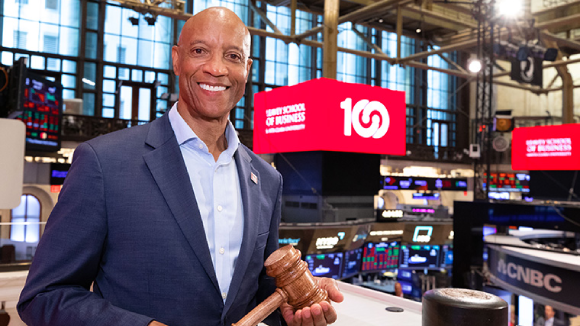 A smiling man holding a gavel at the New York Stock Exchange. Behind him is a red screen with white text reading Leavey School of Business, Santa Clara University, 100