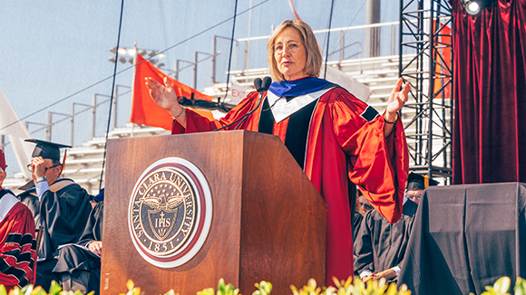 A woman in commencment regalia speaking from a podium with the Santa Clara University seal on the front.