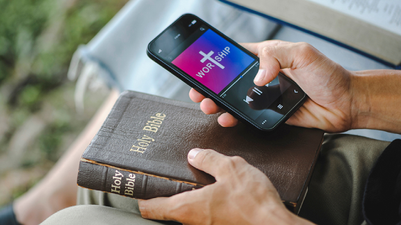 A close-up view of a persons lap. In one hand they hold a smart phone with a graphic that reads Worship with a cross in the middle of the word. In their other hand, they hold a book labeled Holy Bible.