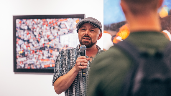 A man standing in front of a wall of paintings. He is speaking into a microphone to a crowd offscreen.