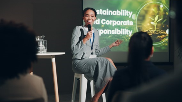 A smiling young woman seated in front of a large display screen. The text on the screen is cut off slightly by her head and torso but reads Sustainability and Corporate Responsibility.