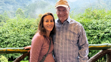 A woman and man, both smiling, standing in front of a green, leafy background. 