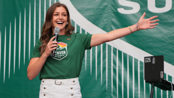 A happy woman holding a microphone and wearing a green shirt with the Denver Summit FC logo, in front of a matching green background.