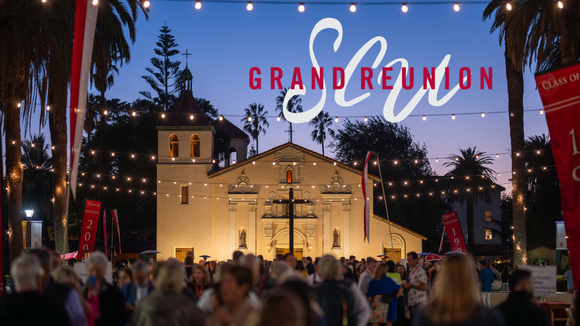 A crowd of people talking and laughing in front of Santa Clara Universitys Mission Church at dusk.