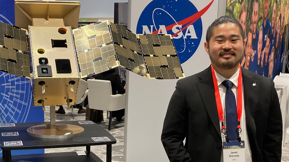 A man wearing a nametag that reads Jared Shimada stands next to a prototype of a probe from NASA.