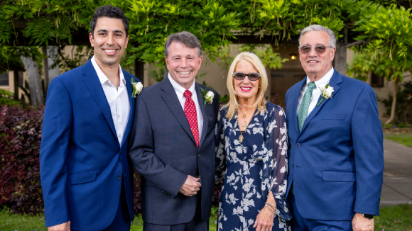 A smiling group of one woman and three men in formal wear, standing in the Santa Clara University Mission Gardens.