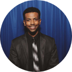 A person in a suit in front of a blue curtain.