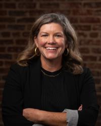 photo of prof megan crane a smiling woman with light brown hair wearing a black shirt and gold necklace and earrings