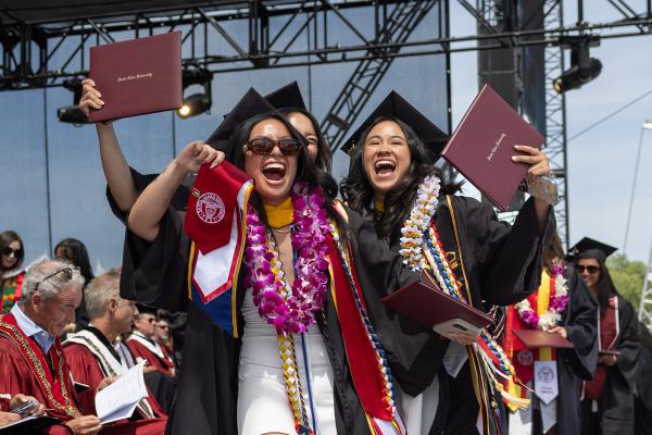 Three students at graduation cheer on stage while holding their diploma up in the air.