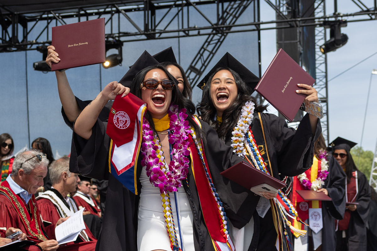 Three students at graduation cheer on stage while holding their diploma up in the air.
