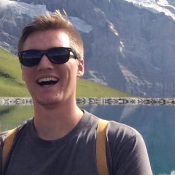 A person smiling outdoors with mountains and a lake in the background.