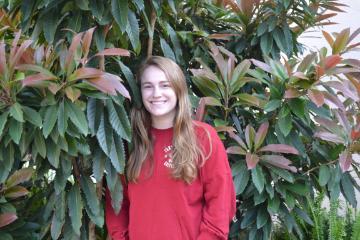 A person in a red sweater standing in front of green shrubs.
