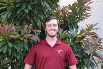 A person in a red polo shirt stands in front of leafy plants.