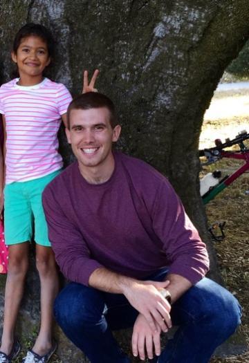 A child standing behind a man crouching and smiling outdoors.