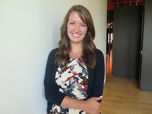 Smiling individual in a floral dress and dark cardigan, standing indoors. image link to story