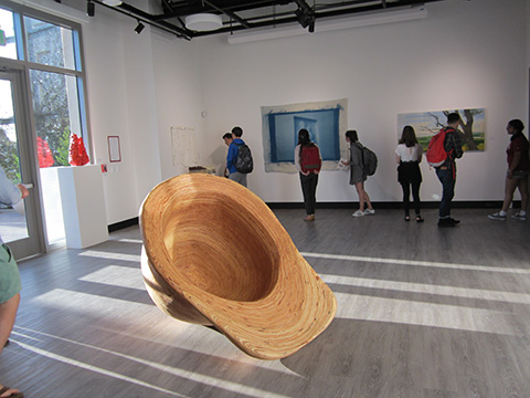 Large hat sculpture displayed in an art gallery with people behind it.