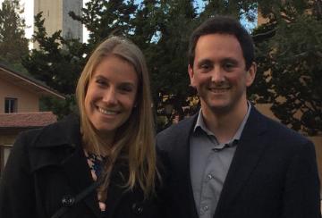 Two people smiling outdoors with buildings and trees in the background. image link to story