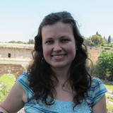 A woman smiling, outdoors, with trees and buildings in the background.