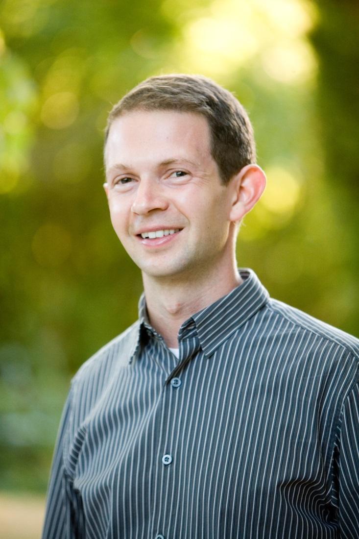 A person smiling outdoors with a blurred green background.