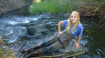 Person sitting on a log in a flowing stream with greenery in the background.