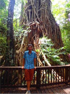 A person posing in front of a large tree within a lush forest.