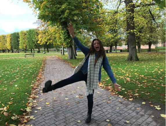 Person posing playfully on a park path with autumn leaves.
