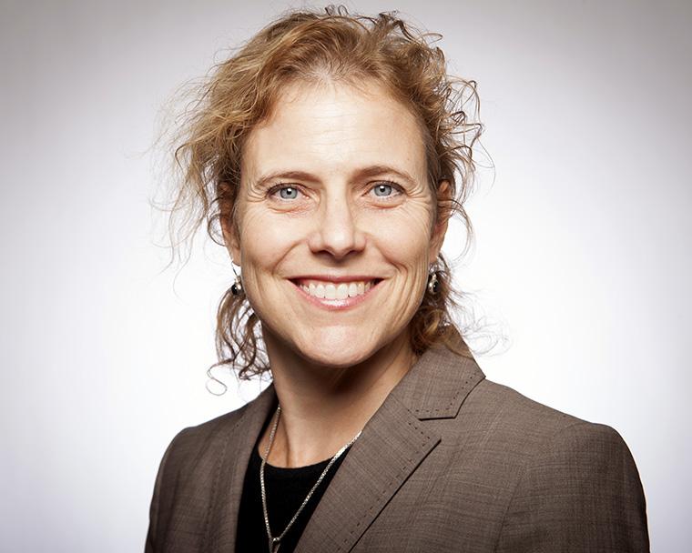 A person in a brown blazer smiling with a neutral background.