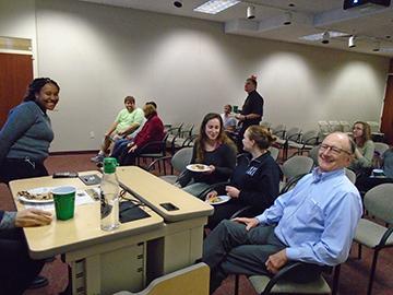 Alt text: People sitting and standing in a room, one person at a table with a laptop. 