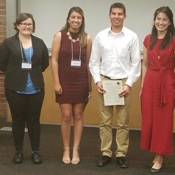 Four people standing in front of a wall; one holding a certificate.