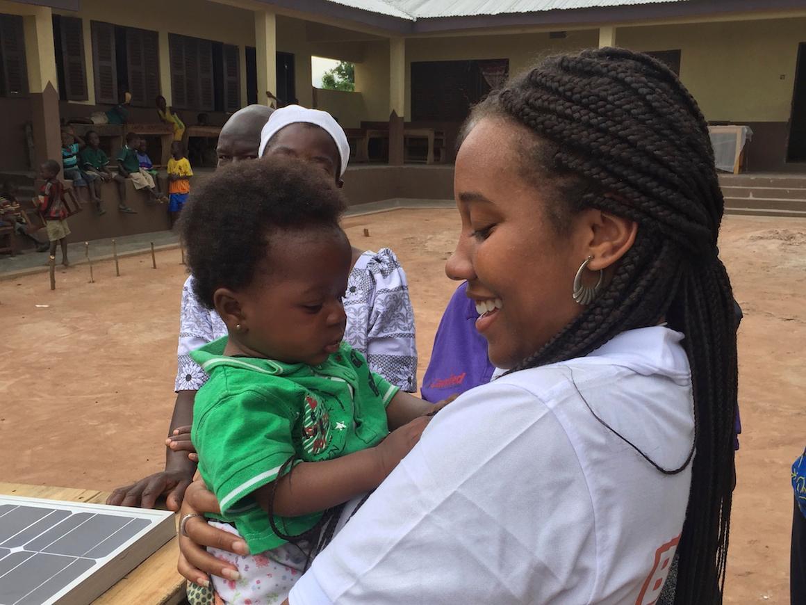 Woman holding a baby outdoors with another person nearby.