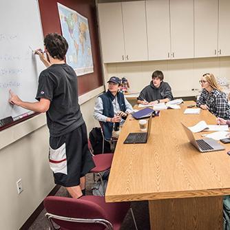 Several students studying at a large table; one writes on a whiteboard. 