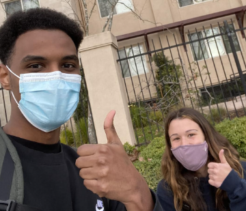 Two masked individuals giving thumbs up outside a community building. image link to story